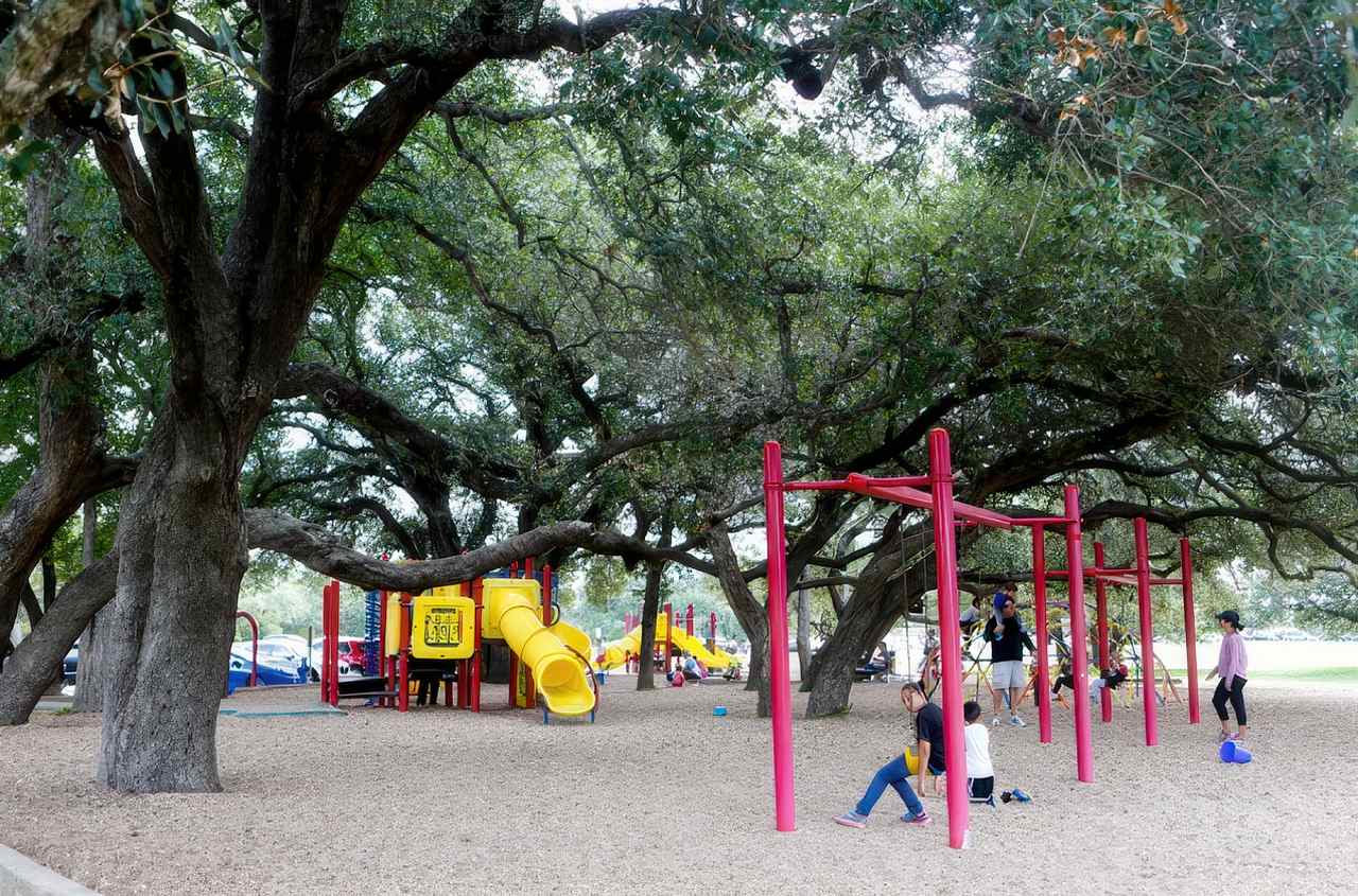 Circle C Metro Park playground features bright red and yellow play structures and swings set beneath a canopy of large, shady oak trees.