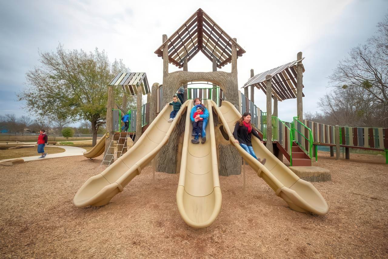 Children slide down wide beige plastic slides built into a large wooden playground structure at Dove Springs Park. The play area features treehouse-style roofs, climbing elements, and a mix of natural and colorful accents, set on a mulch surface surrounded by trees.