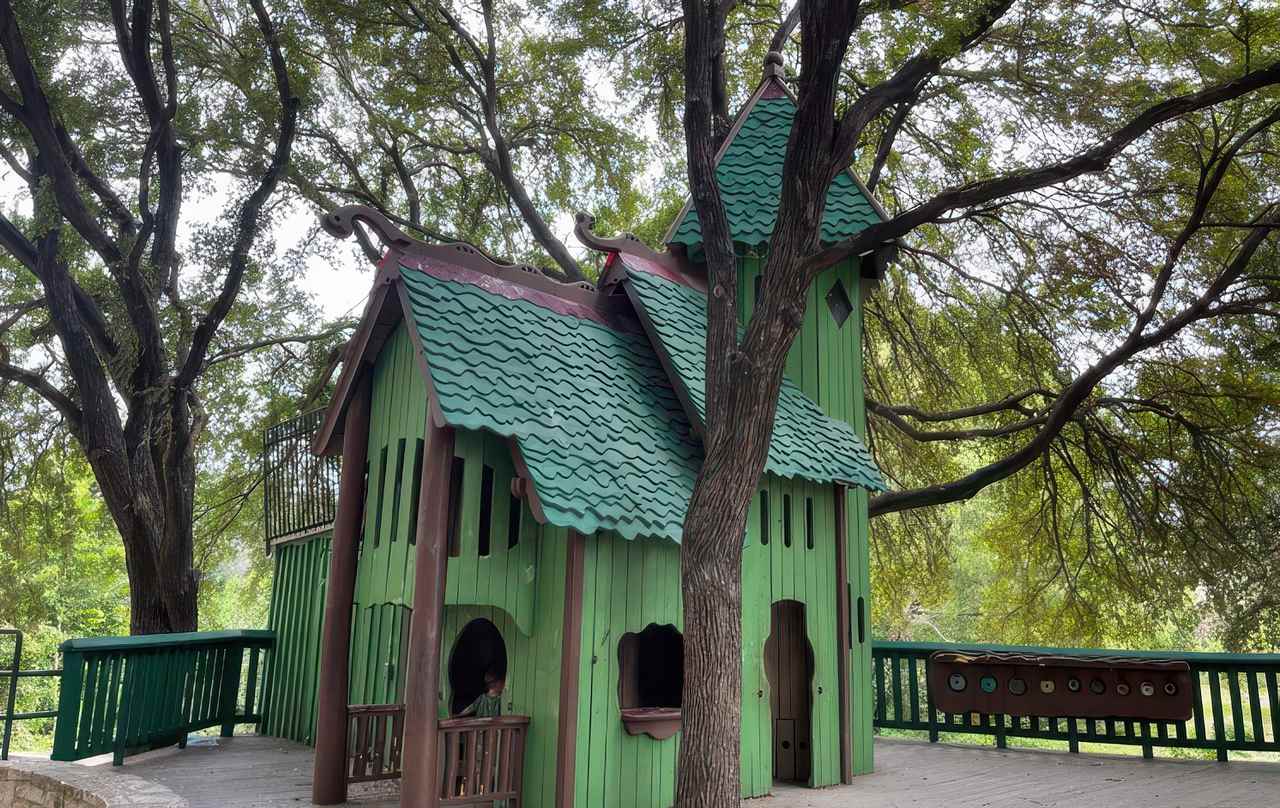 A whimsical green wooden playhouse with wavy rooftops and towers sits under tall trees at Katherine Fleischer Park, inviting imaginative play.