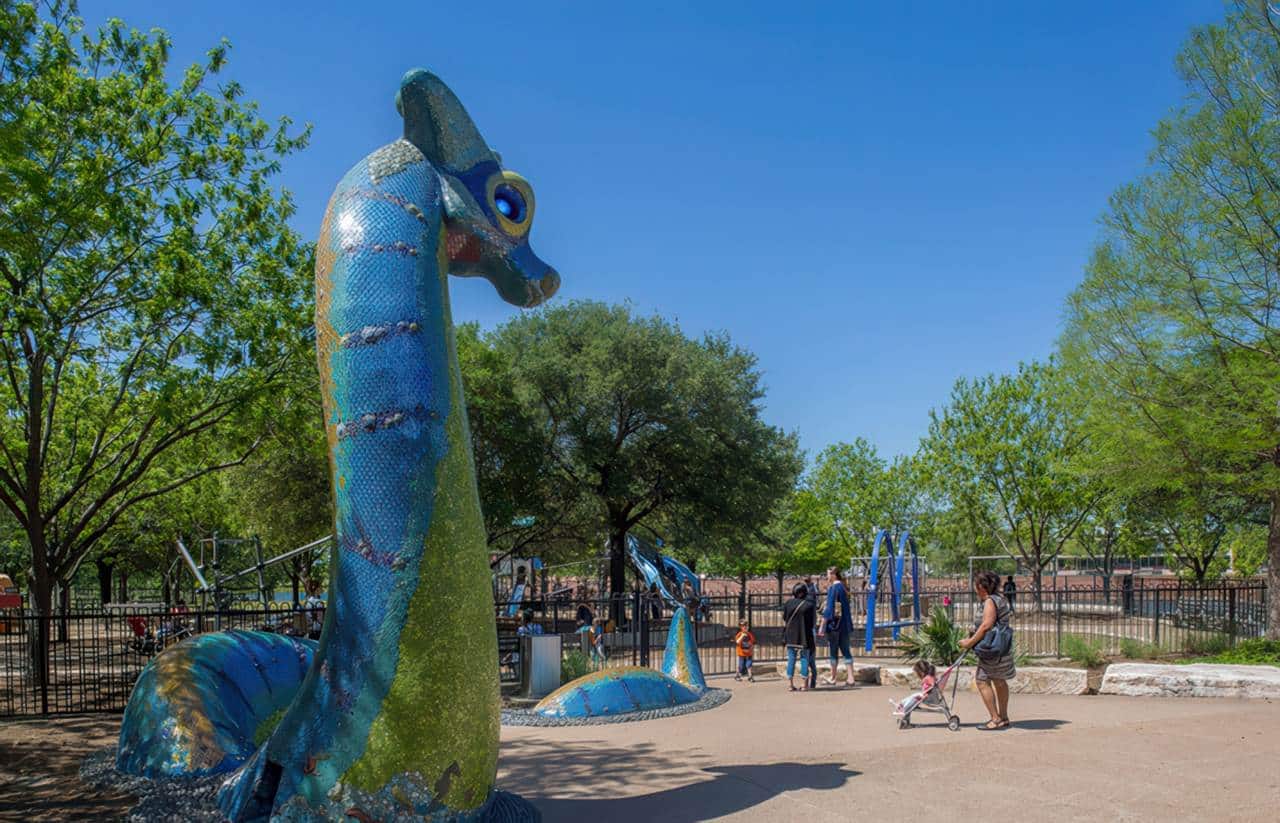 Mueller Park playground features a large blue and green mosaic sea creature sculpture near a fenced play area, with families enjoying a sunny day.