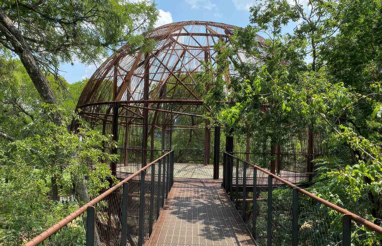 A rust-colored metal dome structure in Pease District Park sits among trees, with an elevated walkway leading to it for climbing and play.