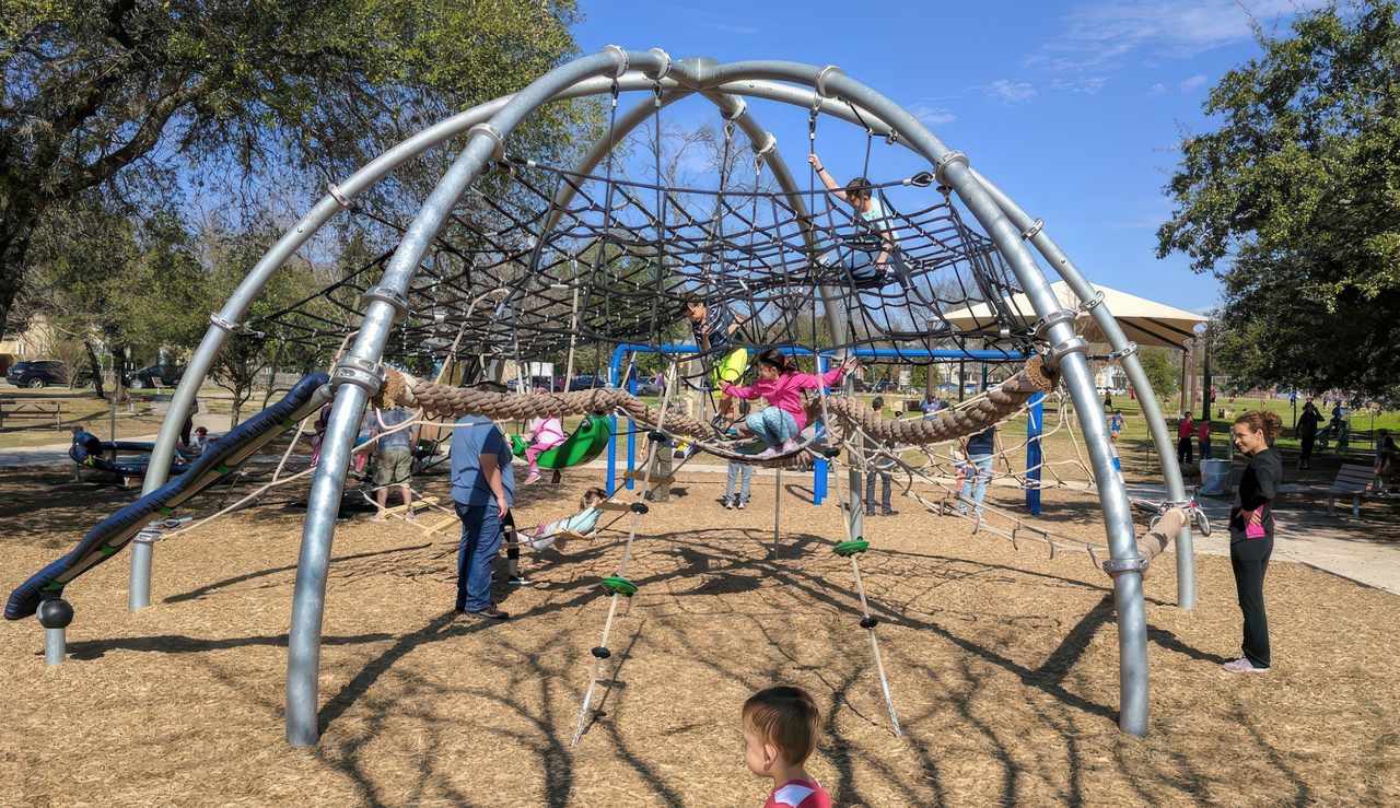 Ramsey Park playground features a large climbing structure shaped like a web dome, where children are seen climbing ropes and nets. The play area is surrounded by trees, wood chip ground cover, and families enjoying the sunny day.