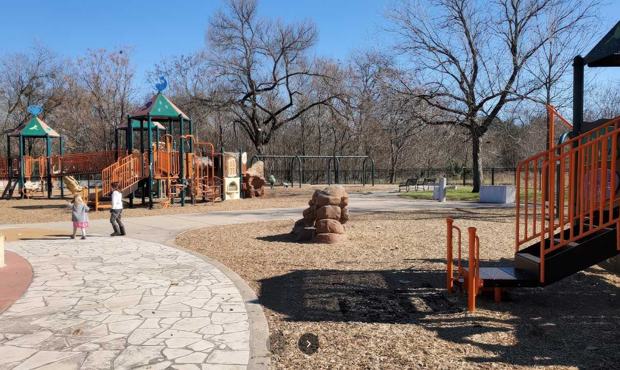 Roy G. Guerrero Park playground features a spacious layout with climbing structures, slides, swings, and natural elements like faux boulders, surrounded by leafless winter trees and open sky.
