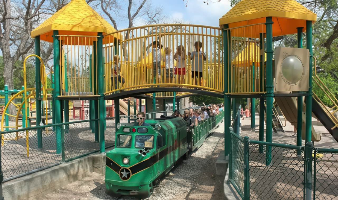 Zilker Park playground features a colorful structure with yellow bridges above a mini train track, where kids play and families ride below.