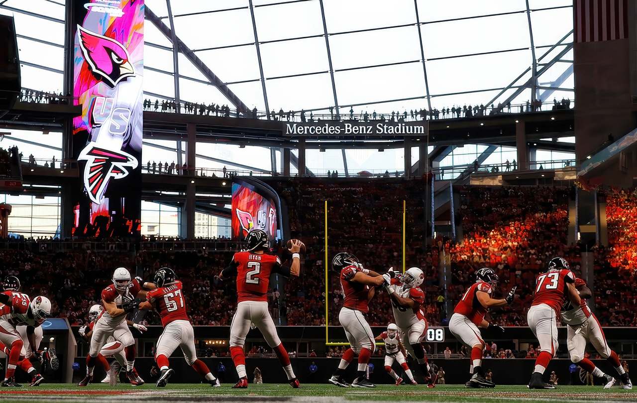 Atlanta Falcons quarterback Matt Ryan prepares to throw a pass during a game at Mercedes-Benz Stadium, with teammates blocking Arizona Cardinals defenders. The stadium's retractable roof is open, letting in daylight as fans fill the stands.