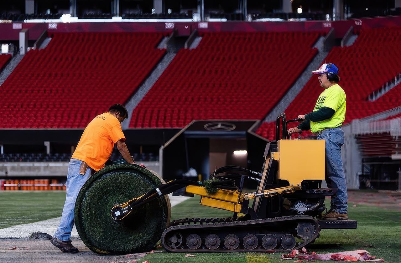 Two workers installing artificial turf inside a stadium using a yellow machine, with rolls of synthetic grass and red seating visible in the background, demonstrating field preparation or turf maintenance.