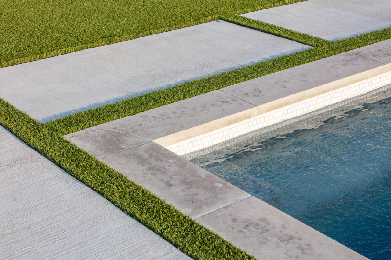 A modern poolside design with clean concrete pavers separated by strips of artificial grass, creating a sharp geometric layout.