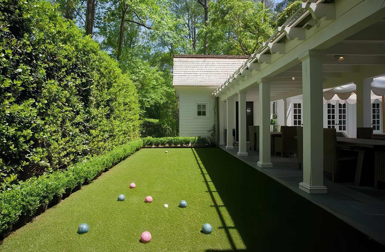 A private backyard bocce ball court with artificial grass lies beside a white house and a covered patio, surrounded by tall green hedges.