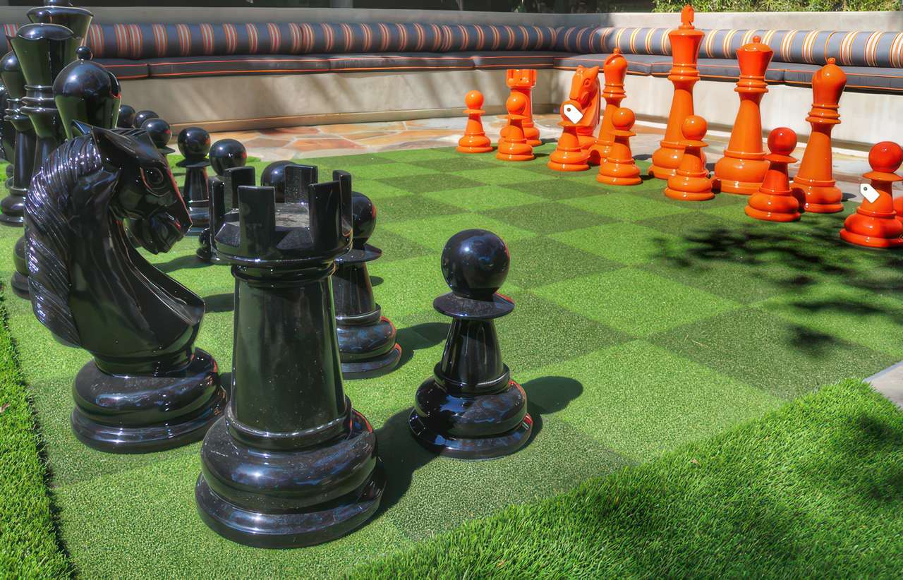 A large outdoor chessboard made on artificial grass features oversized black and orange chess pieces near a cushioned seating area.