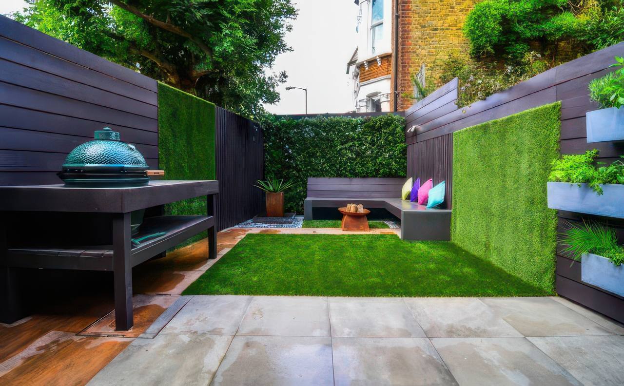 A cozy urban garden entertainment area features artificial grass, vertical greenery, colorful pillows on bench seating, and a modern grill.