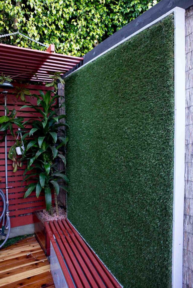 A small garden corner with a vertical artificial grass wall, red bench seating, wood deck flooring, and tropical plants under a slatted canopy.
