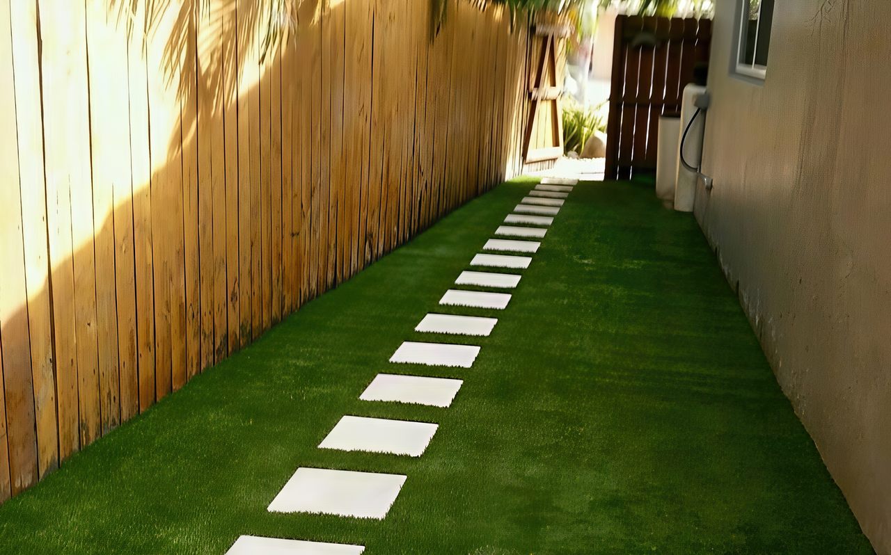 A narrow side yard features artificial grass with a straight row of white stepping stones between a wooden fence and house wall.