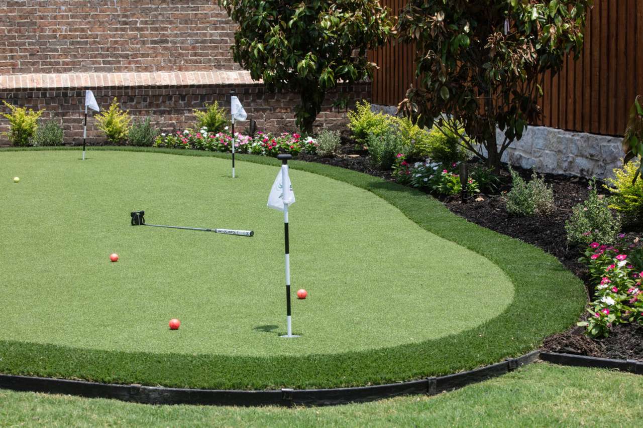 A backyard putting green with red golf balls, a putter, and four small flags, bordered by flowers and brick and wood fencing.