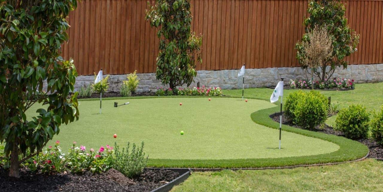 A backyard putting green at a residential home in Texas, surrounded by flags, shrubs, and colorful flower beds.