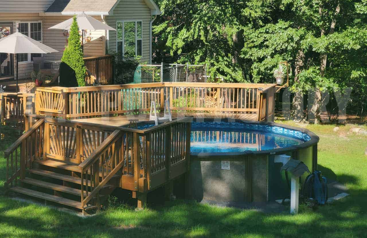 Above-ground pool with surrounding wooden deck, steps, and patio umbrellas, set in a grassy backyard near a wooded area.