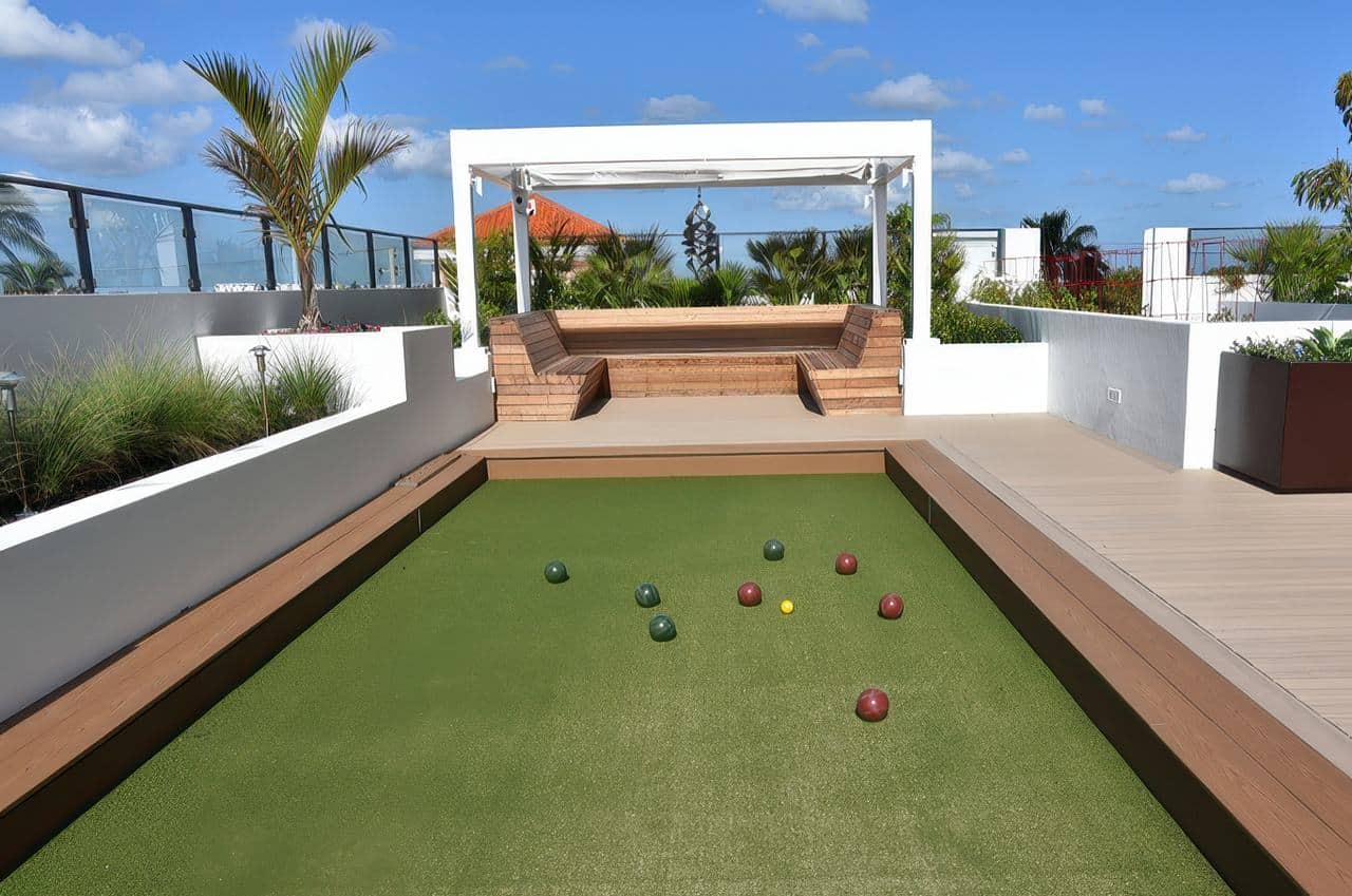 Modern rooftop bocce ball court with artificial turf, surrounding benches, and a shaded wooden seating area under a clear blue sky.