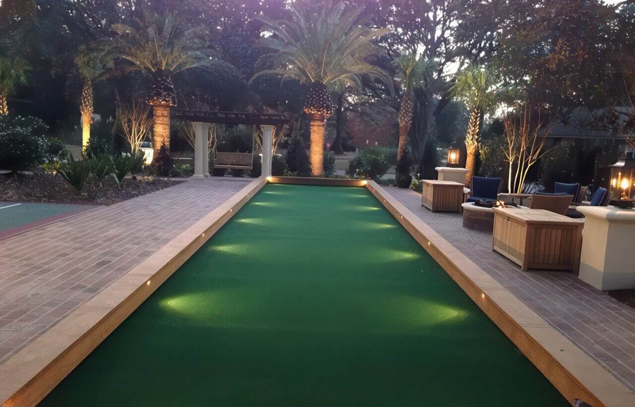 Elegant backyard bocce ball court with artificial turf, accent lighting, palm trees, and cozy seating areas under evening lights.