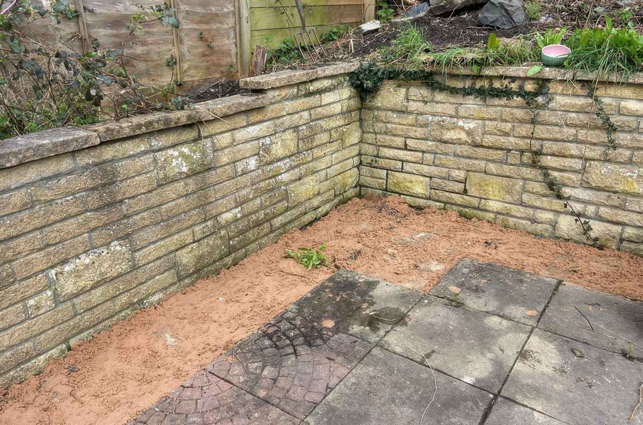 Small concrete backyard with aged paving stones and a narrow strip of bare soil along a stone retaining wall. The wall has patches of moss and climbing ivy, and some garden tools and plants are visible at the top edge.