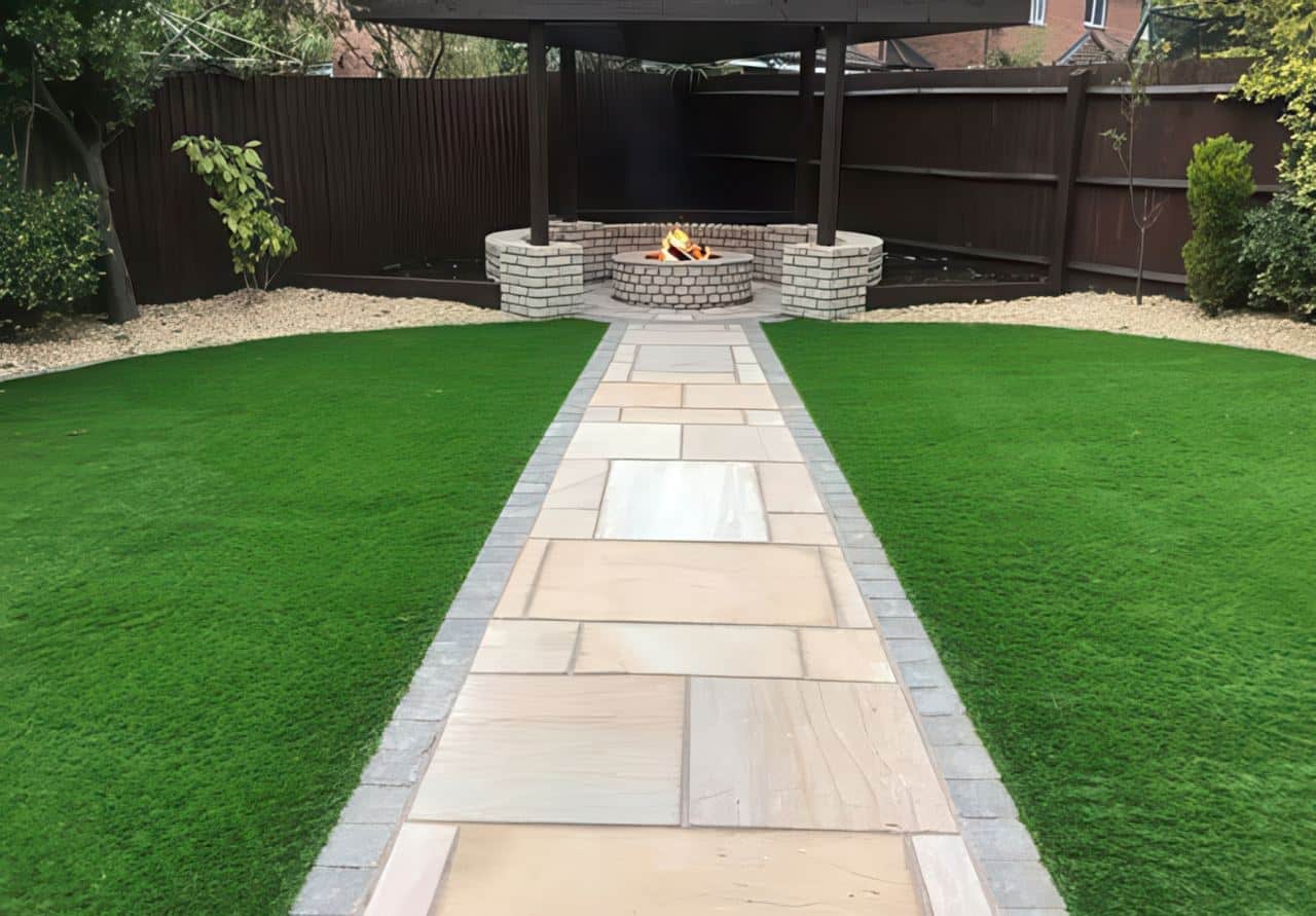 Backyard with a stone pathway flanked by artificial grass, leading to a circular brick firepit under a dark wood pergola.