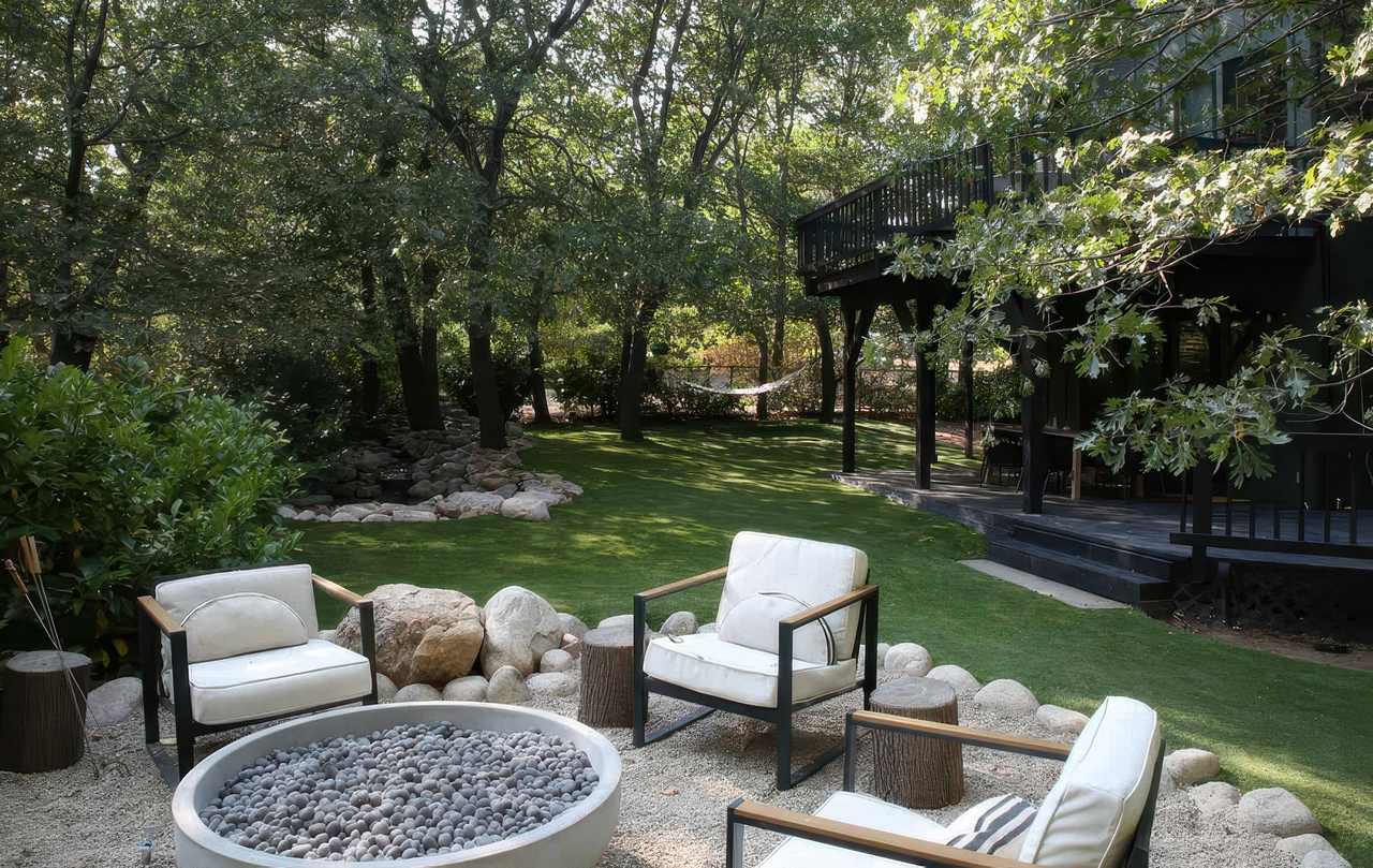 Peaceful backyard with pea gravel firepit area, white cushioned chairs, artificial grass lawn, mature trees, and a black elevated deck.