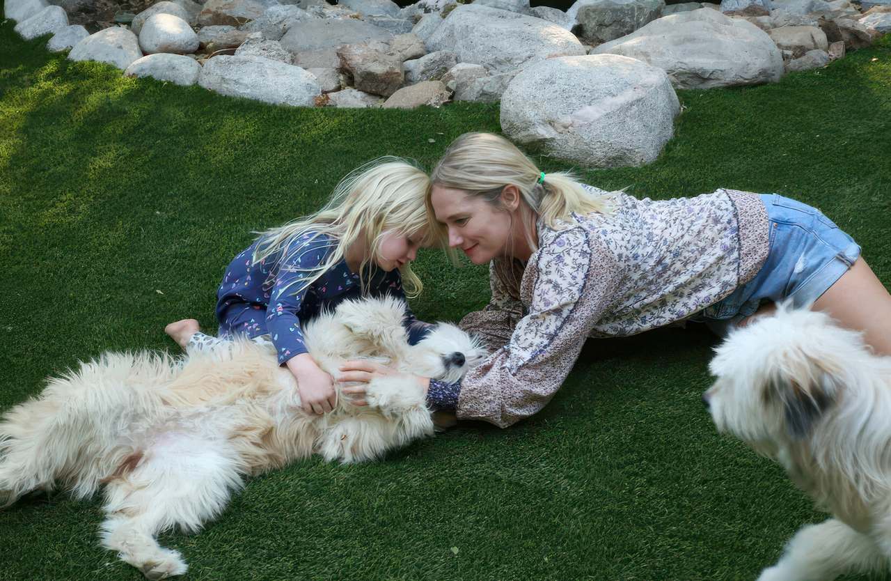 Mother and daughter lying on artificial grass, cuddling a fluffy dog near a rock border in a backyard, with another dog approaching.