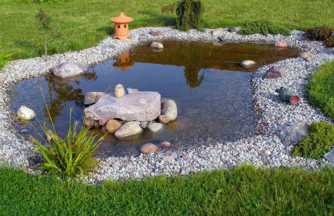 Small decorative pond surrounded by smooth pebbles, rocks, grass, and a terracotta lantern, with clear water and natural landscaping.
