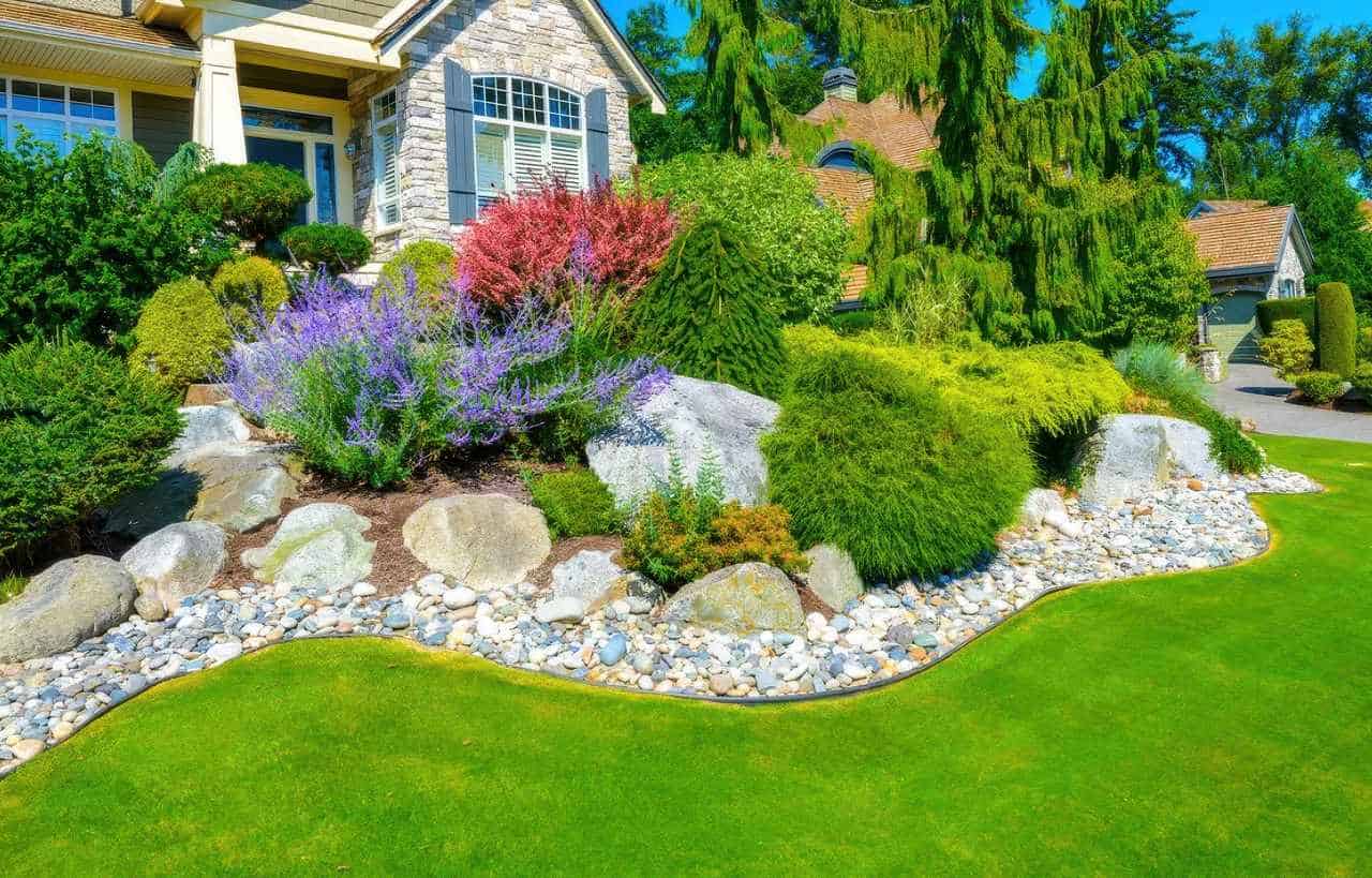 Vibrant front yard with artificial grass, a colorful rock garden featuring boulders, shrubs, and flowering plants near a stone house.
