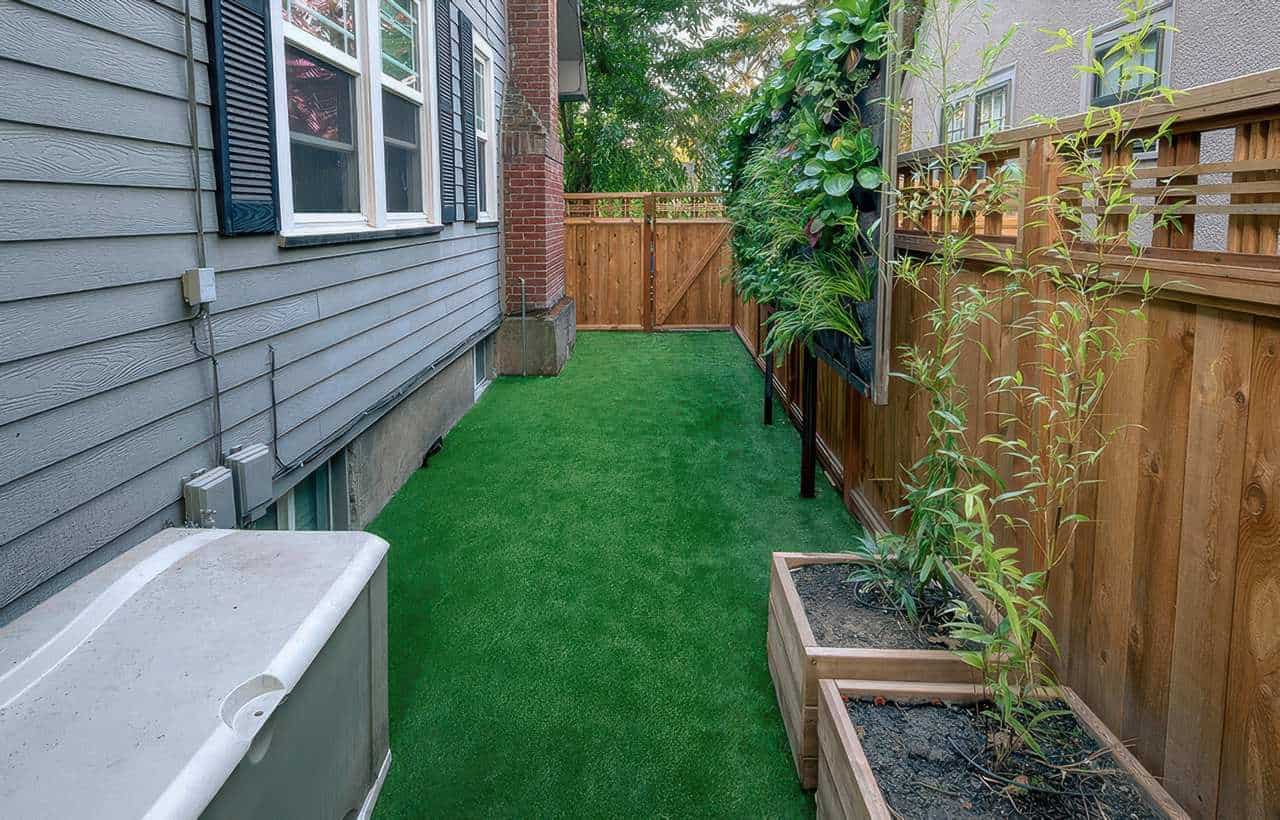Narrow side yard with artificial grass, bordered by a wooden fence with vertical planters and raised wooden planter boxes containing young plants, next to a gray house with black shutters and utility boxes.