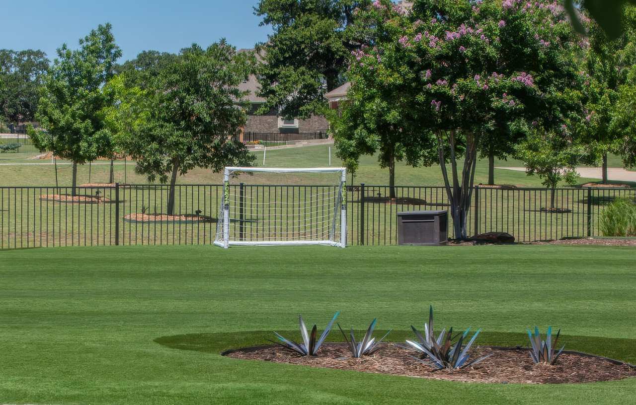 Spacious backyard with artificial grass, a small soccer goal, metal fencing, and landscaped trees and plants under a clear blue sky.