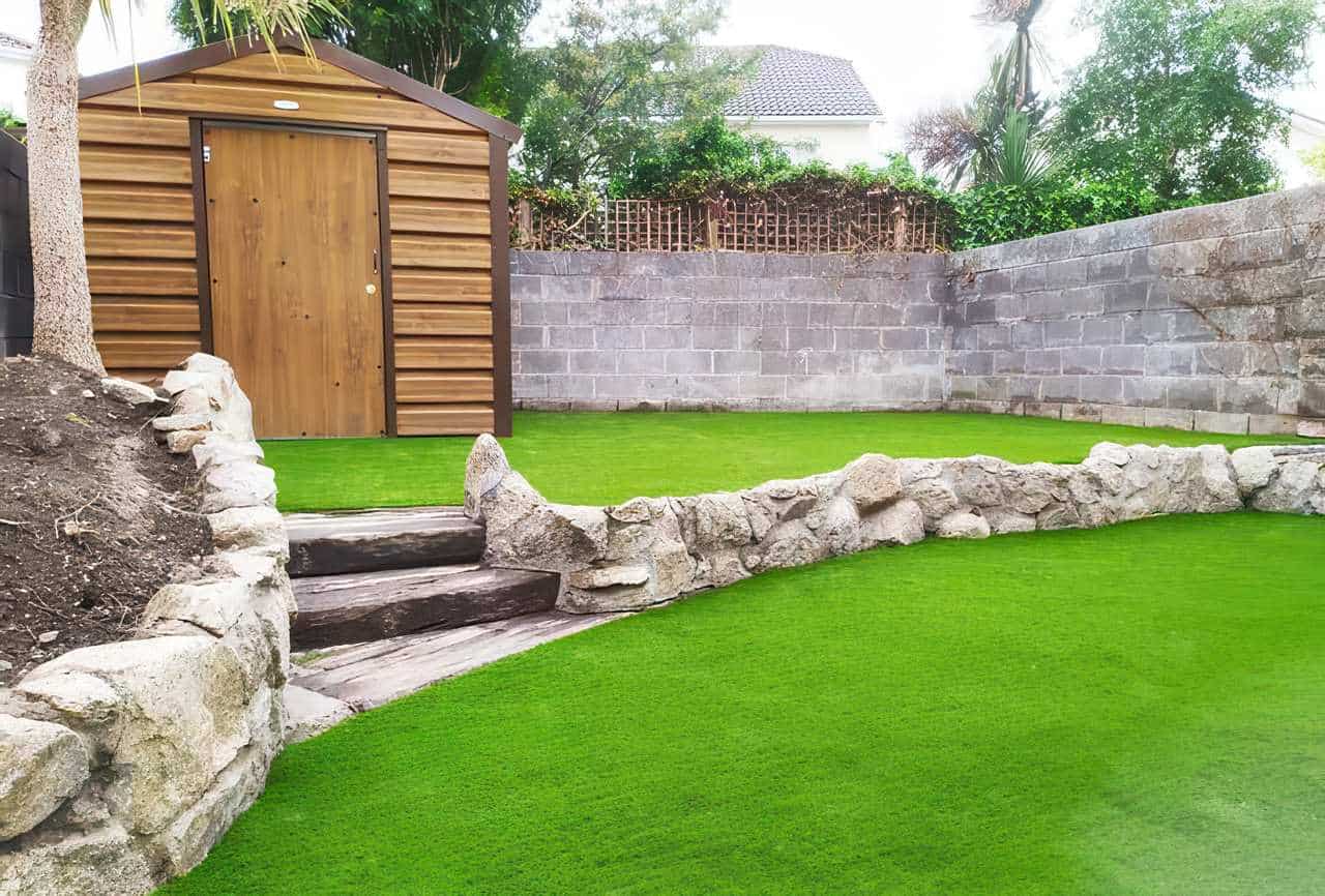 Backyard transformed with bright artificial grass covering the ground, bordered by rustic stone edging and wooden steps leading to a higher section. A modern wooden shed stands against a gray block wall, with trees and greenery in the background.