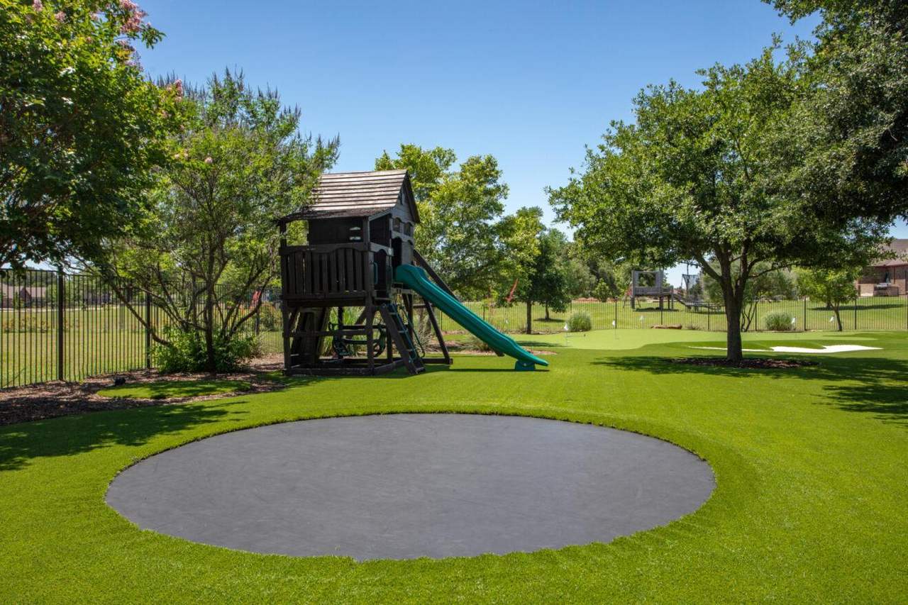 Spacious backyard with artificial grass, featuring an in-ground trampoline, wooden playhouse with slides, and shaded trees near a fenced lawn.