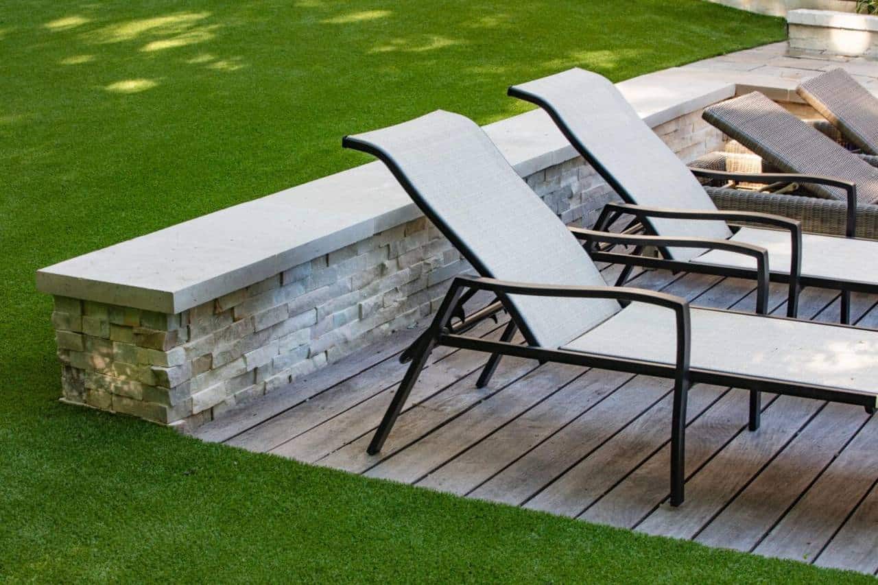 Artificial grass installed around a patio with lounge chairs and stone seating wall, creating a modern, low-maintenance backyard.