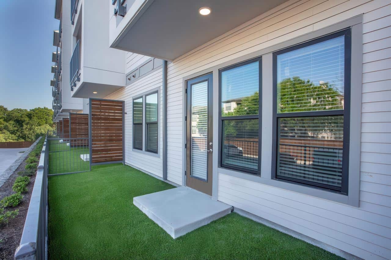 Apartment patio with artificial grass, fenced yard, wood privacy screens, and modern white siding with large windows.