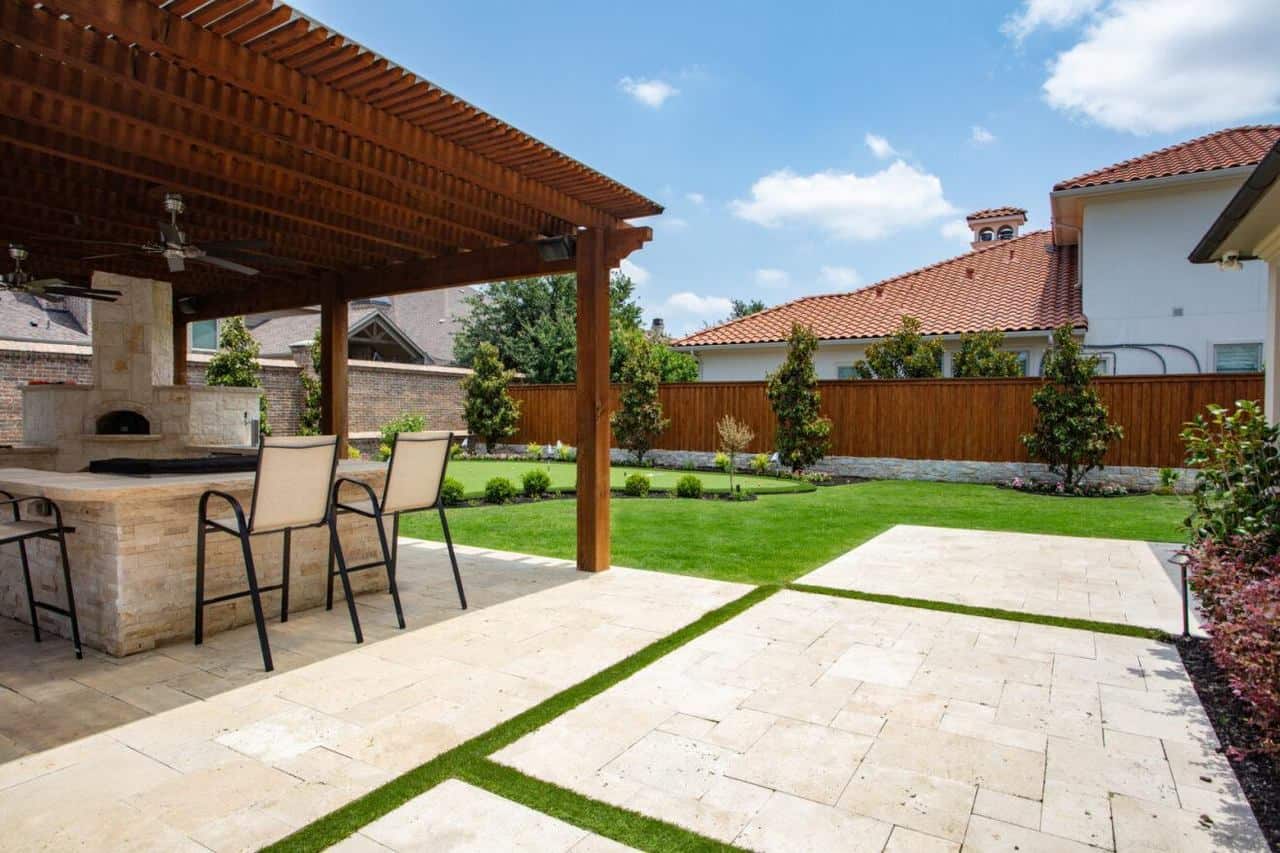 Backyard with a covered outdoor kitchen and bar, stone pavers with turf strips, and a manicured lawn bordered by young trees.