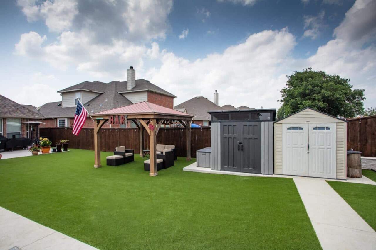 Backyard with artificial grass, patio seating under a pergola labeled "BAR," and two storage sheds beside a concrete path.