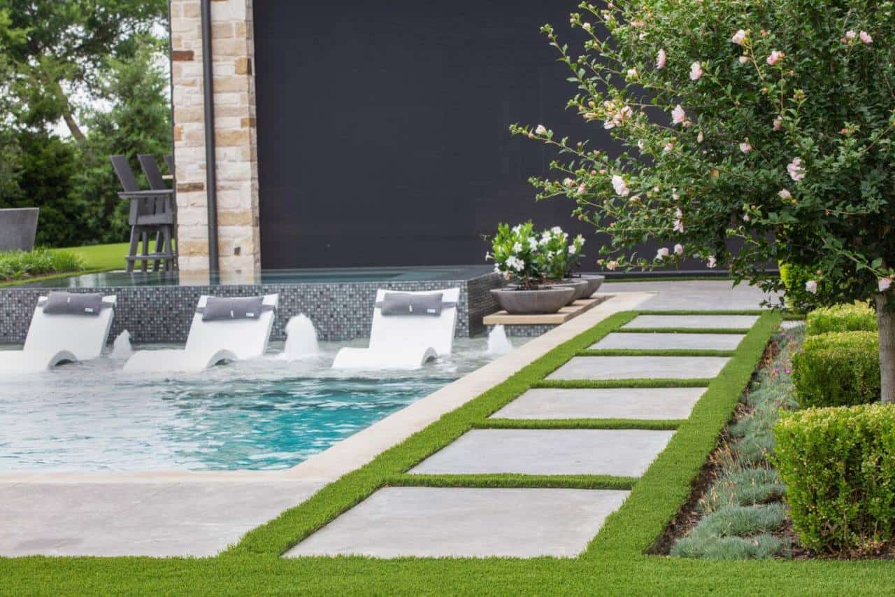 Modern backyard pool with in-water lounge chairs, concrete pavers set in turf, and neatly trimmed landscaping with flowers.