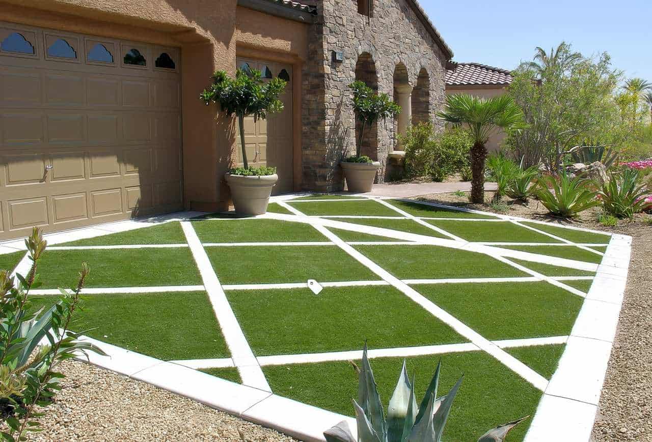 Desert-style driveway featuring bold white pavers in a crisscross pattern set in artificial grass, beside a stone and stucco home.