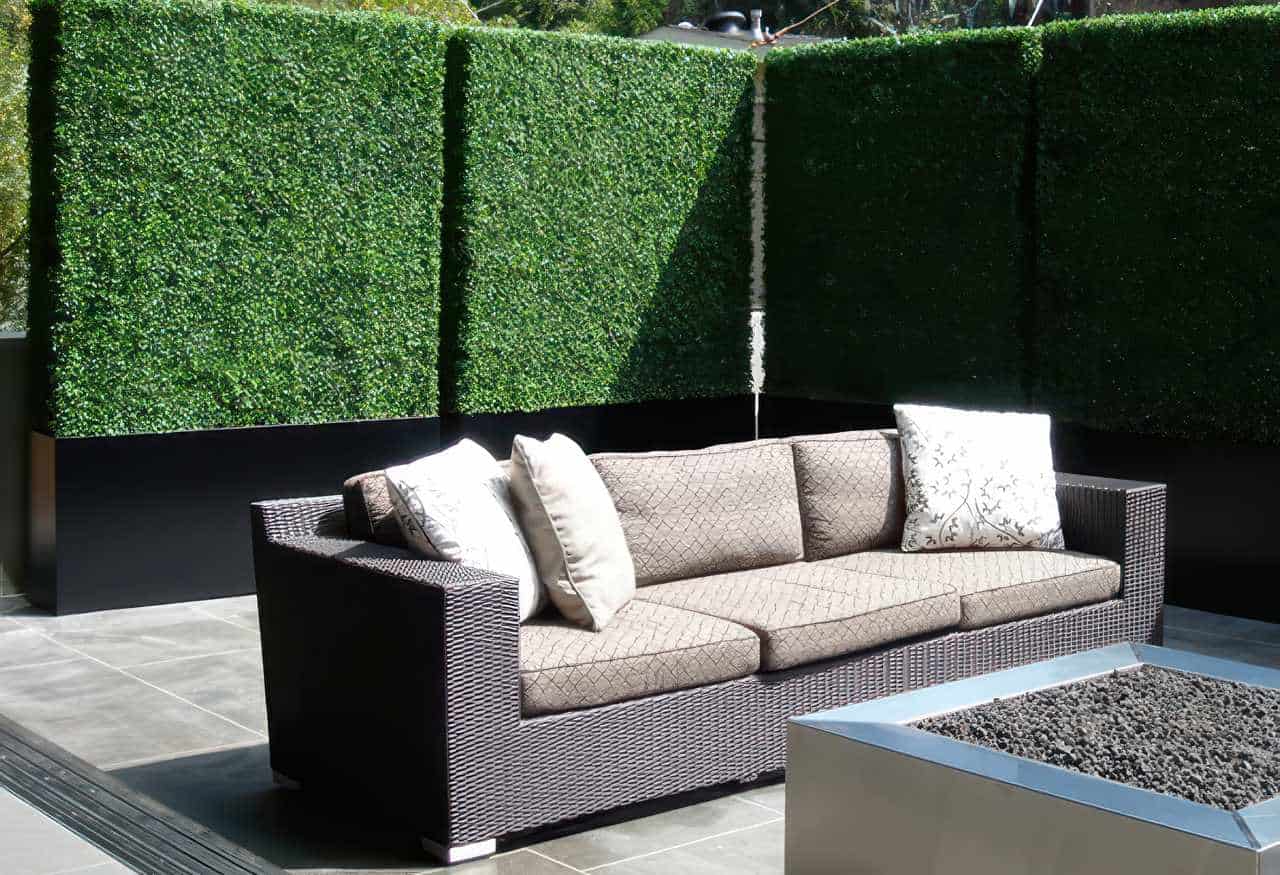 Modern small backyard with a dark wicker outdoor sofa, beige cushions, decorative pillows, and artificial green privacy hedge panels behind it, complemented by a sleek square fire pit filled with black stones on a tiled patio floor.