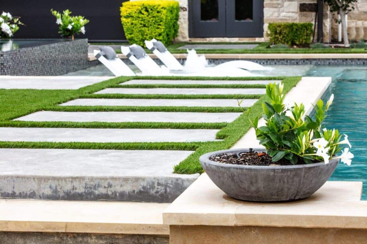 Contemporary poolside with turf-lined concrete pavers, in-pool loungers, and a round planter with blooming white flowers.