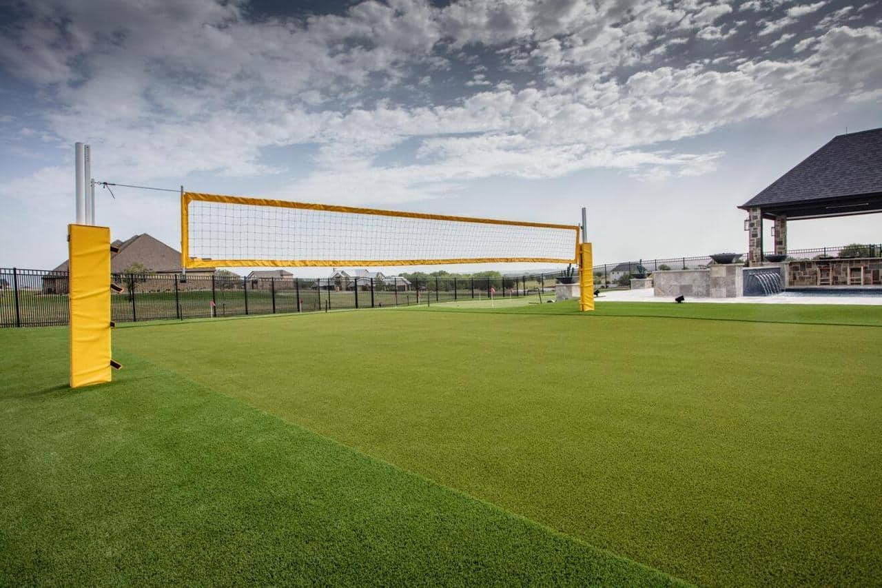 Backyard volleyball court with artificial turf, yellow net posts, and a nearby covered patio with a pool and waterfall feature.