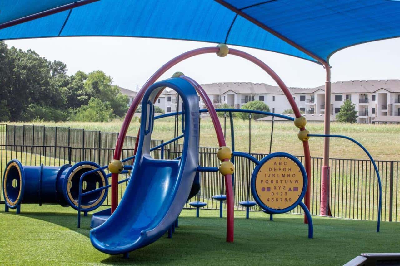 Colorful playground with blue slides, tunnels, alphabet play panel, and arched climbing bars under a large blue sunshade on artificial grass.